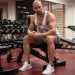 Strong, determined man in white tank top sitting in gym, showcasing fitness and strength.